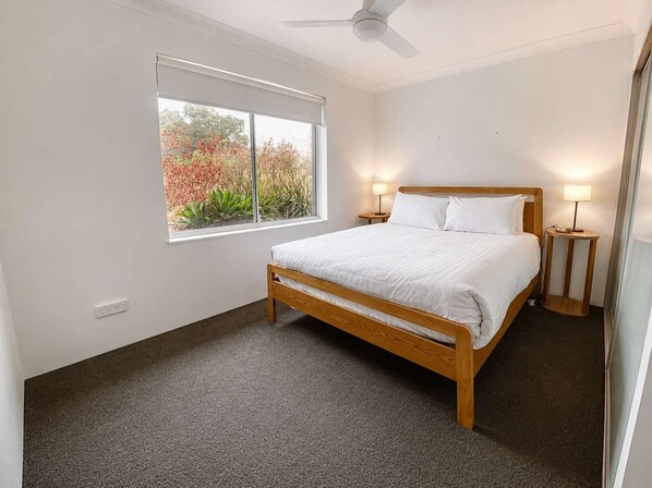 A bedroom with a single bed, a ceiling fan, a window with a view of foliage, and two bedside lamps.