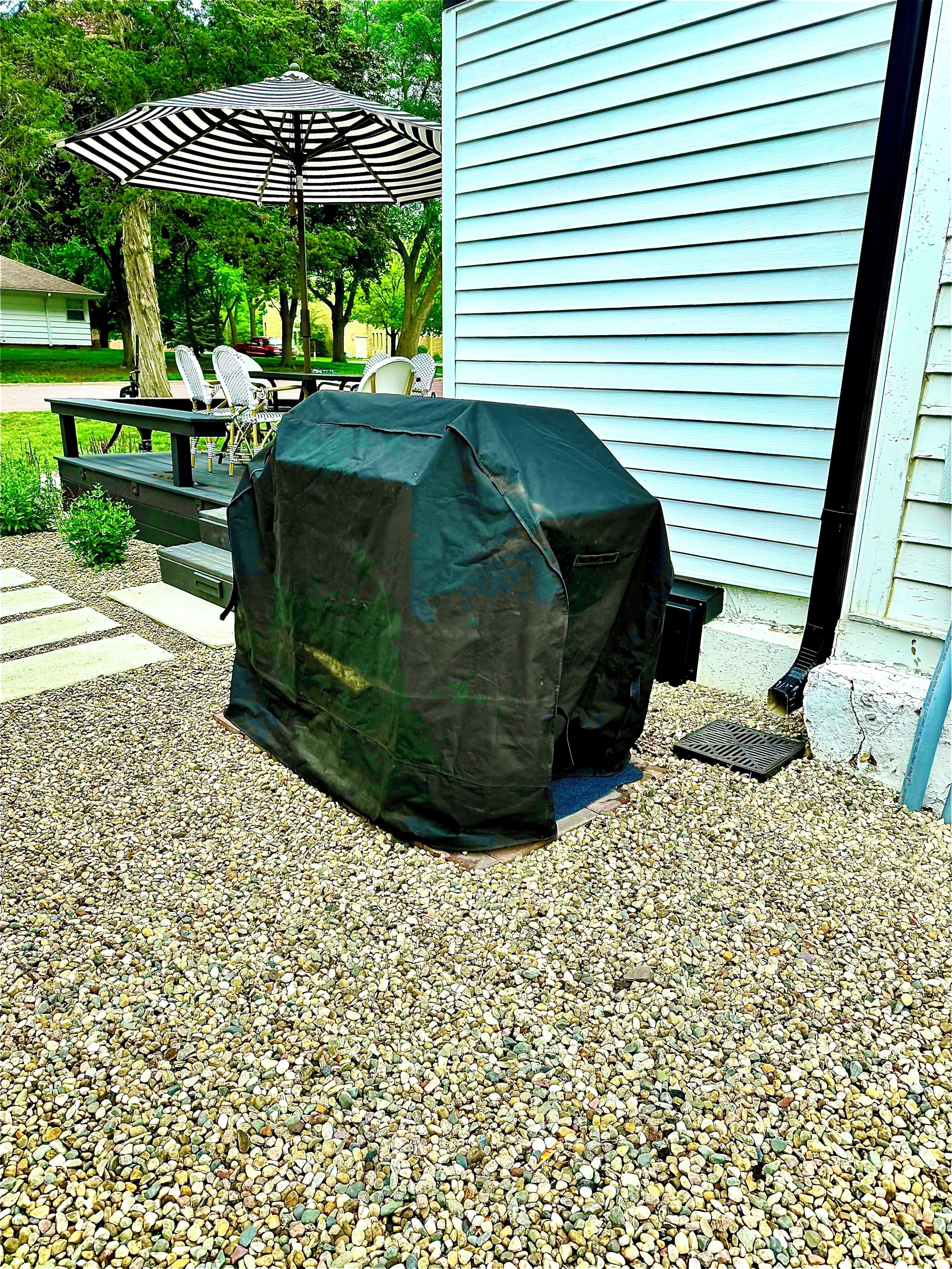 Un barbecue recouvert d’une bâche noire de protection, installé sur une terrasse en gravier avec une plateforme en bois et des meubles d’extérieur en arrière-plan.