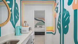 A modern bathroom with a white sink, a mirror, and a toilet. There is a white cabinet with a microwave and a teal coffee maker on the counter. The walls are decorated with abstract leaf patterns in teal, orange, and yellow. - Royal Palms Motel (Tybee Island)