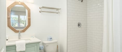 Un baño con lavamanos blanco, un espejo, inodoro y zona de ducha con azulejos blancos.