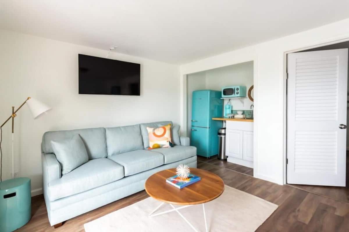Ein modernes Wohnzimmer mit einem hellblauen Sofa, einem runden Couchtisch mit einem Buch und einer Dekoration, einem flachbildfernseher an der Wand und einer Küchenzeile mit einem türkisfarbenen Kühlschrank und weißen Schränken.