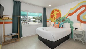 A modern hotel room with a bed, a window with curtains, a wall with a colorful mural, and a small table with a lamp. - Royal Palms Motel (Tybee Island)