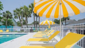 A pool area with yellow lounge chairs and striped umbrellas, surrounded by a white fence and palm trees. - Royal Palms Motel (Tybee Island)