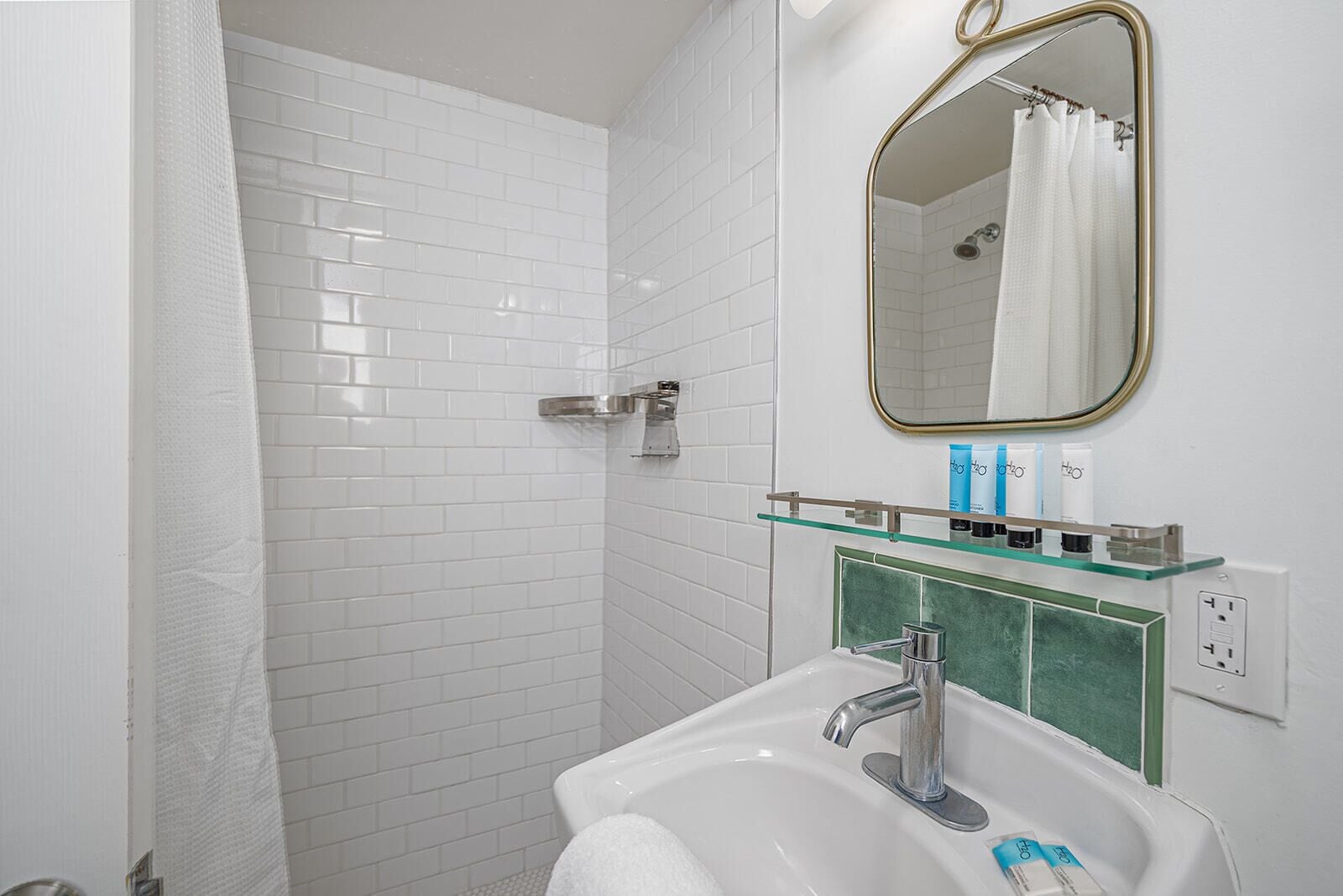 Un baño con paredes de azulejos blancos, estante de vidrio con artículos de aseo, lavamanos blanco y zona de ducha.