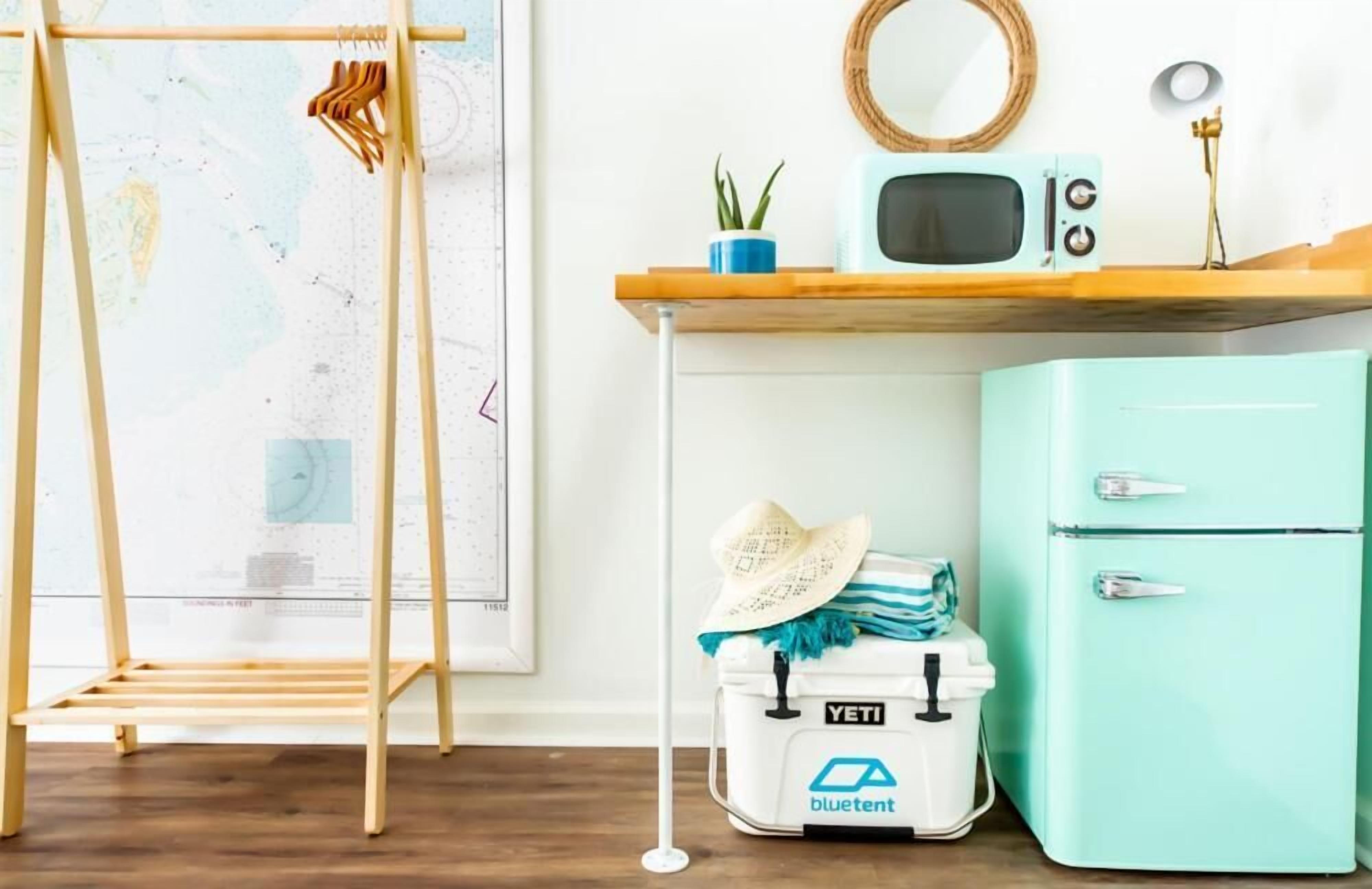 A room with a wooden ladder, a white cooler with a blue tent logo, a mint green refrigerator, and a wooden shelf with a small plant and a vintage radio.