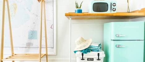 A room with a wooden ladder, a white cooler with a blue tent logo, a mint green refrigerator, and a wooden shelf with a small plant and a vintage radio.