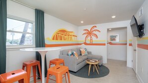A modern hotel room with a grey sofa, orange stools, a small round table, and a TV mounted on the wall. - Royal Palms Motel (Tybee Island)