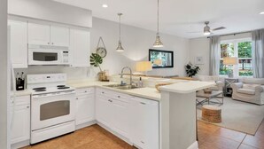 A modern kitchen with white cabinets, a built-in microwave, and a stove. There is a dining area with a white table and chairs, and a living room with a sofa and a TV.