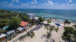 Une vue aérienne d’une propriété située en bord de mer, avec une jetée s’étendant dans des eaux turquoise, une plage de sable dotée de chaises longues et un petit bâtiment surmonté d’un toit rouge.