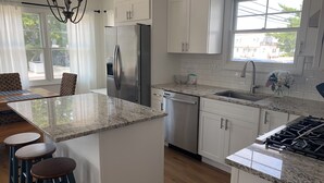 A modern kitchen with white cabinets, a stainless steel refrigerator, a dishwasher, and a granite countertop. There are three stools and a window with a view of the outside.