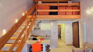 Ein modernes Interieur mit einer Holztreppe zu einem Schlafzimmer-Loft, einer Küchenzeile und einer Sitzecke mit orangefarbenen Stühlen.