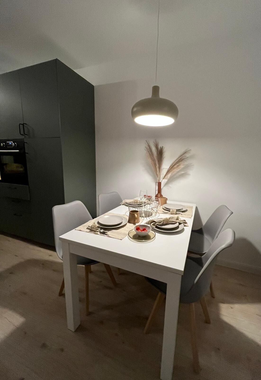Un comedor con una mesa blanca dispuesta para cuatro personas, rodeada de sillas grises. Una lámpara colgante moderna ilumina la mesa.