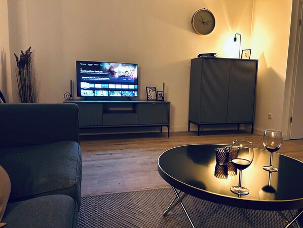 Un salon avec un téléviseur à écran plat posé sur un meuble, un canapé foncé, une table basse ronde en verre avec des verres à vin, et un meuble adossé au mur.