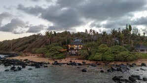 Una casa costera con techo oscuro, rodeada de abundante vegetación y palmeras, con vistas a una costa rocosa.