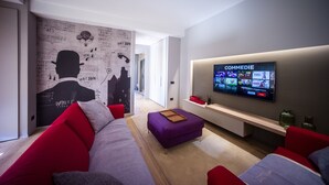 Ett modernt vardagsrum med en stor platt-TV som visar en strömnings­tjänst, en röd och grå soffa, en lila pall och en vägg med en väggmålning som föreställer en stadsbild.