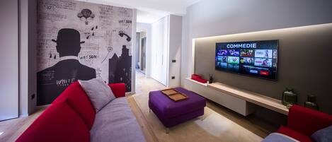 A modern living room with a large flat-screen TV displaying a streaming service, a red and grey sofa, a purple ottoman, and a wall with a cityscape mural.