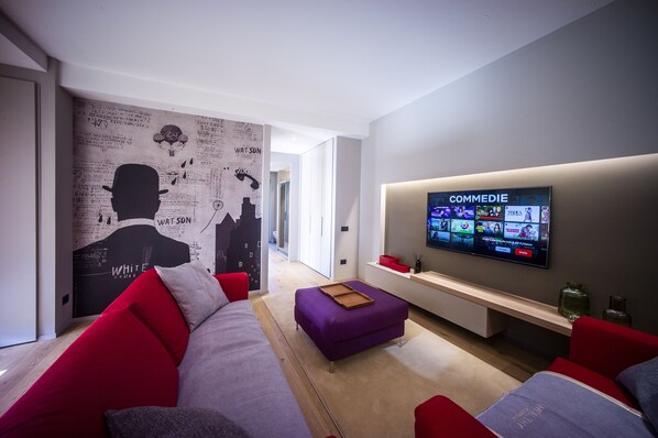 A modern living room with a large flat-screen TV displaying a streaming service, a red and grey sofa, a purple ottoman, and a wall with a cityscape mural.