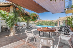An outdoor patio with a pool, stone flooring, white plastic chairs, and a table with fruit.