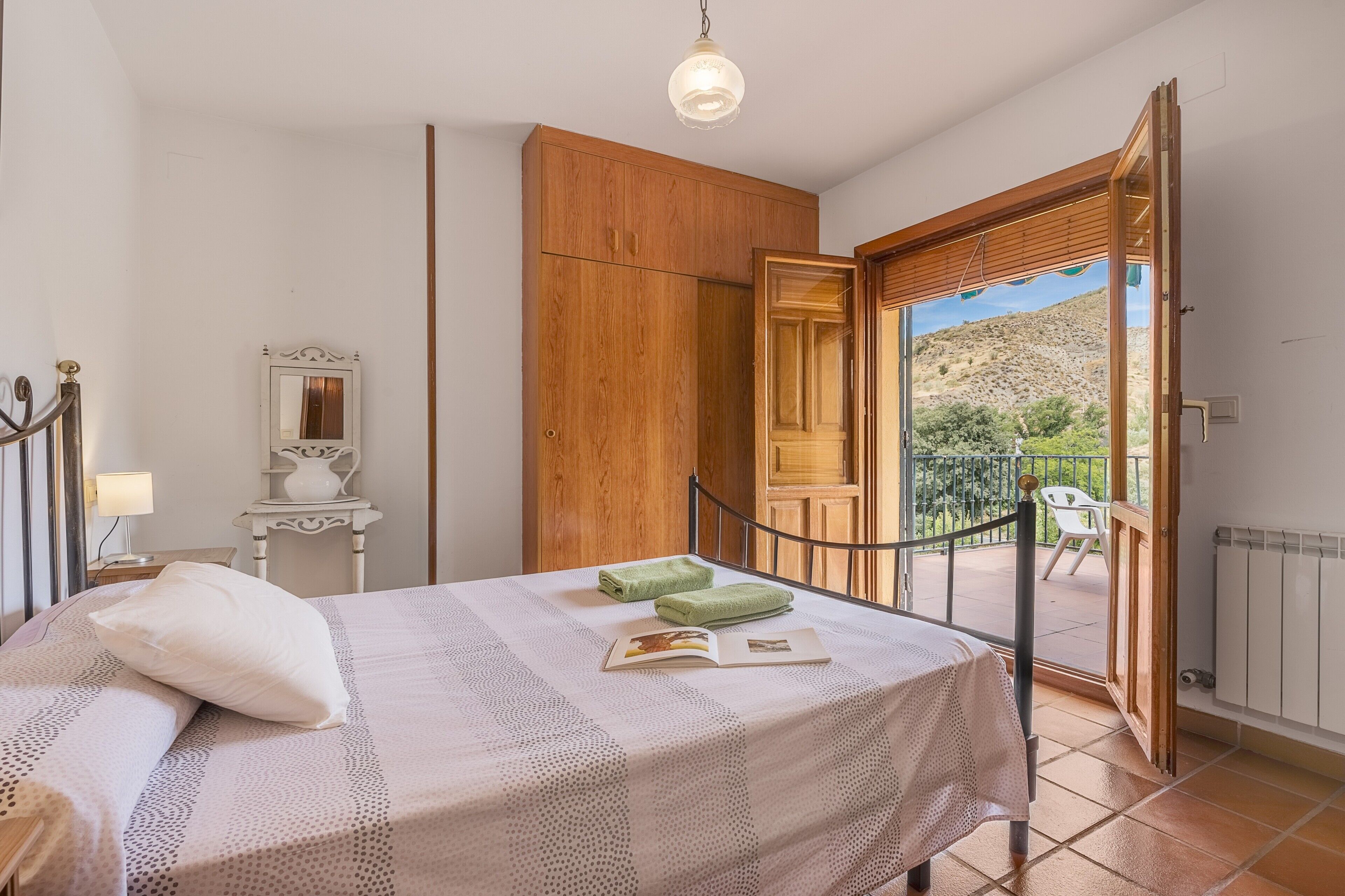 A bedroom with a bed, a nightstand, a mirror, a wooden wardrobe, and a balcony with a view of the hills.