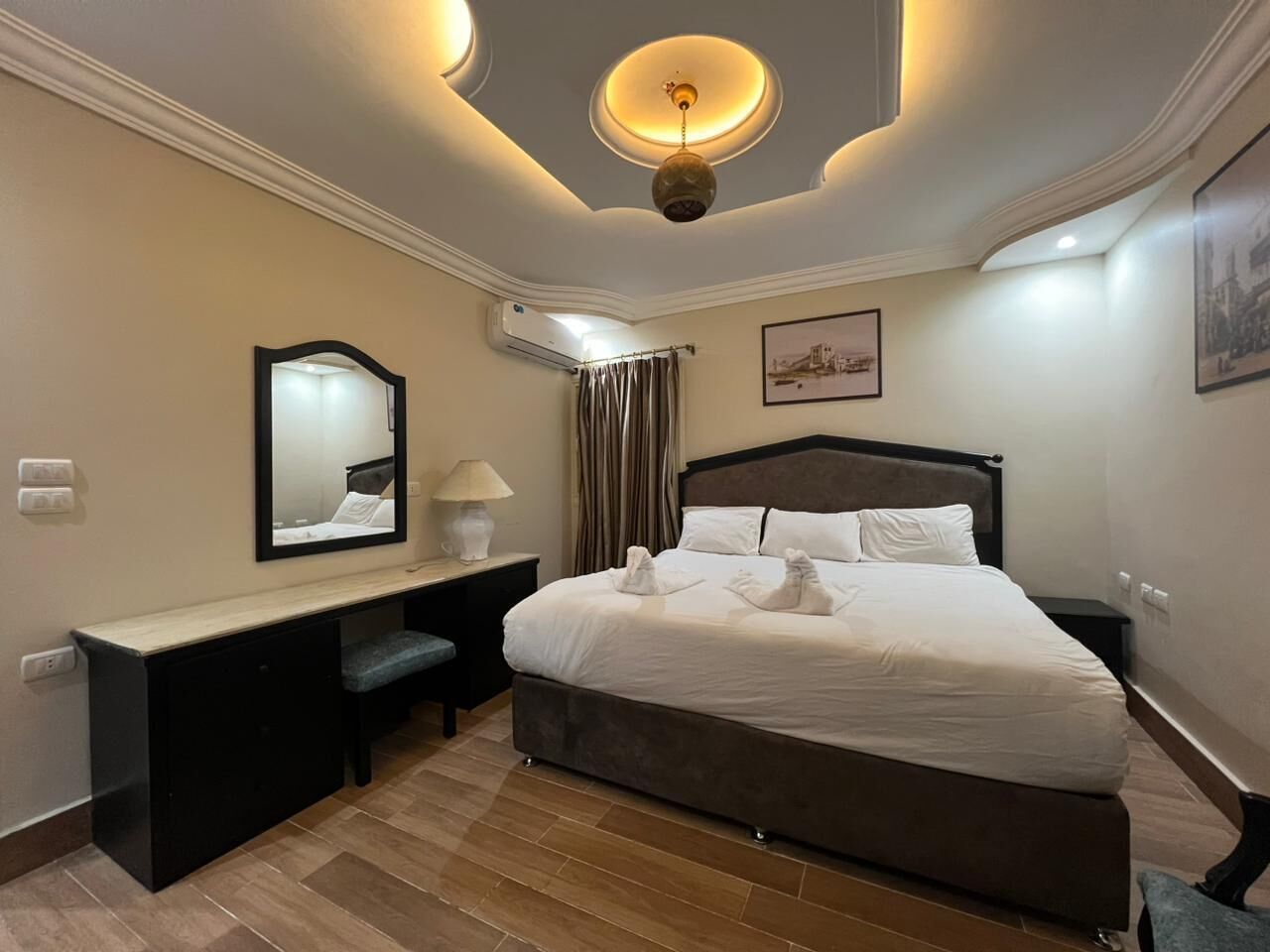 A hotel room with a large bed, a desk with a chair, a mirror, a lamp, and a ceiling light fixture.