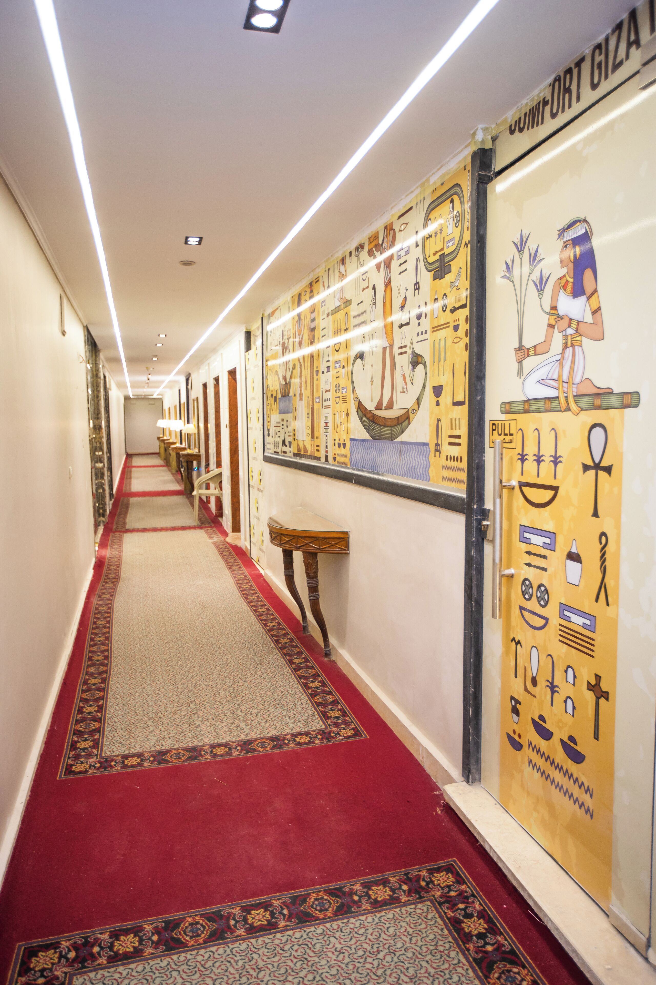 Ein Flur mit rotem Teppich, Wandbildern im ägyptischen Stil und einem Schild mit der Aufschrift 'COMFORT GIZA'.