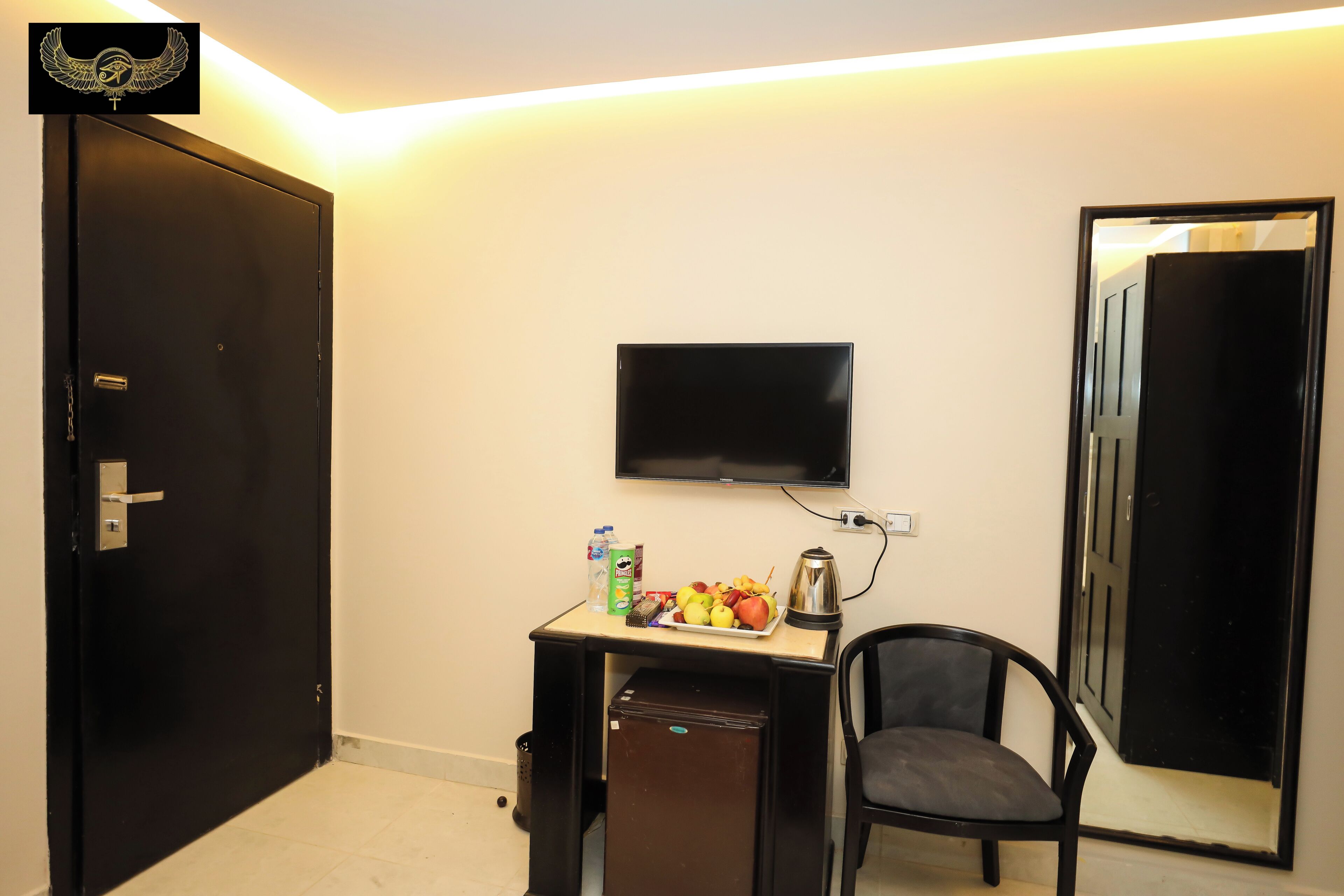 A compact room with a black door, a small desk with a flat-screen TV, a chair, and a refrigerator.