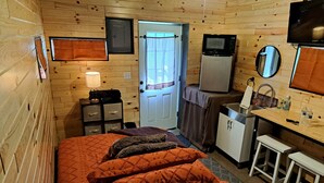 Quarto compacto com cama, escrivaninha, geladeira, televisão e espelho.