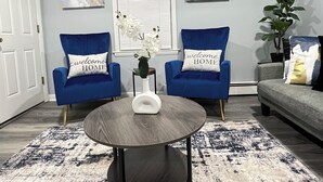 A living room with two blue armchairs, a round coffee table, and a gray sofa. There are two abstract paintings on the wall, a white door, and a window with blinds. - Cozy 2-bedroom apartment in charming New Britain with WiFi (New Britain)