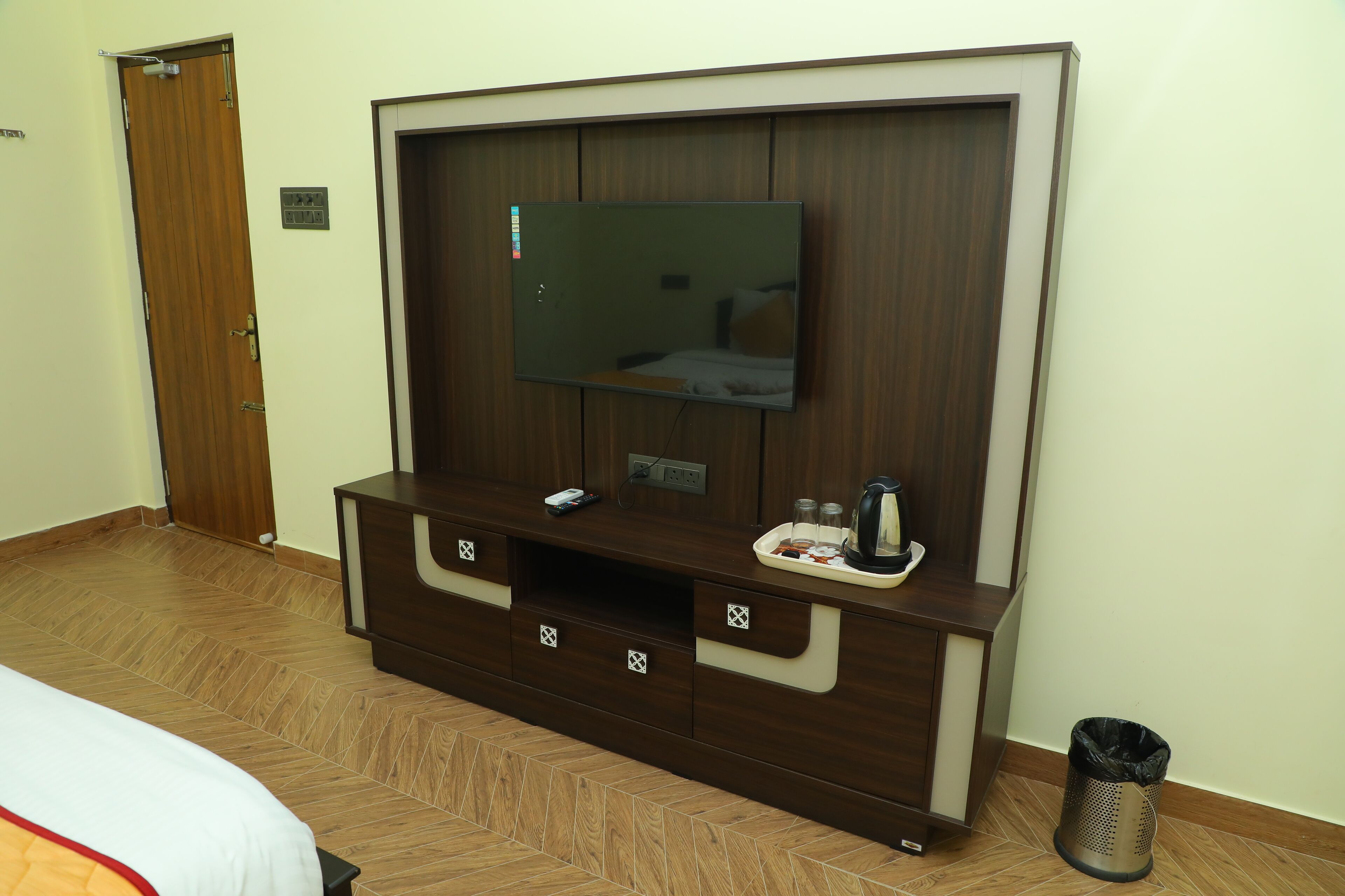 A hotel room with a wooden cabinet, a flat-screen TV, a bed, a trash bin, and a door.