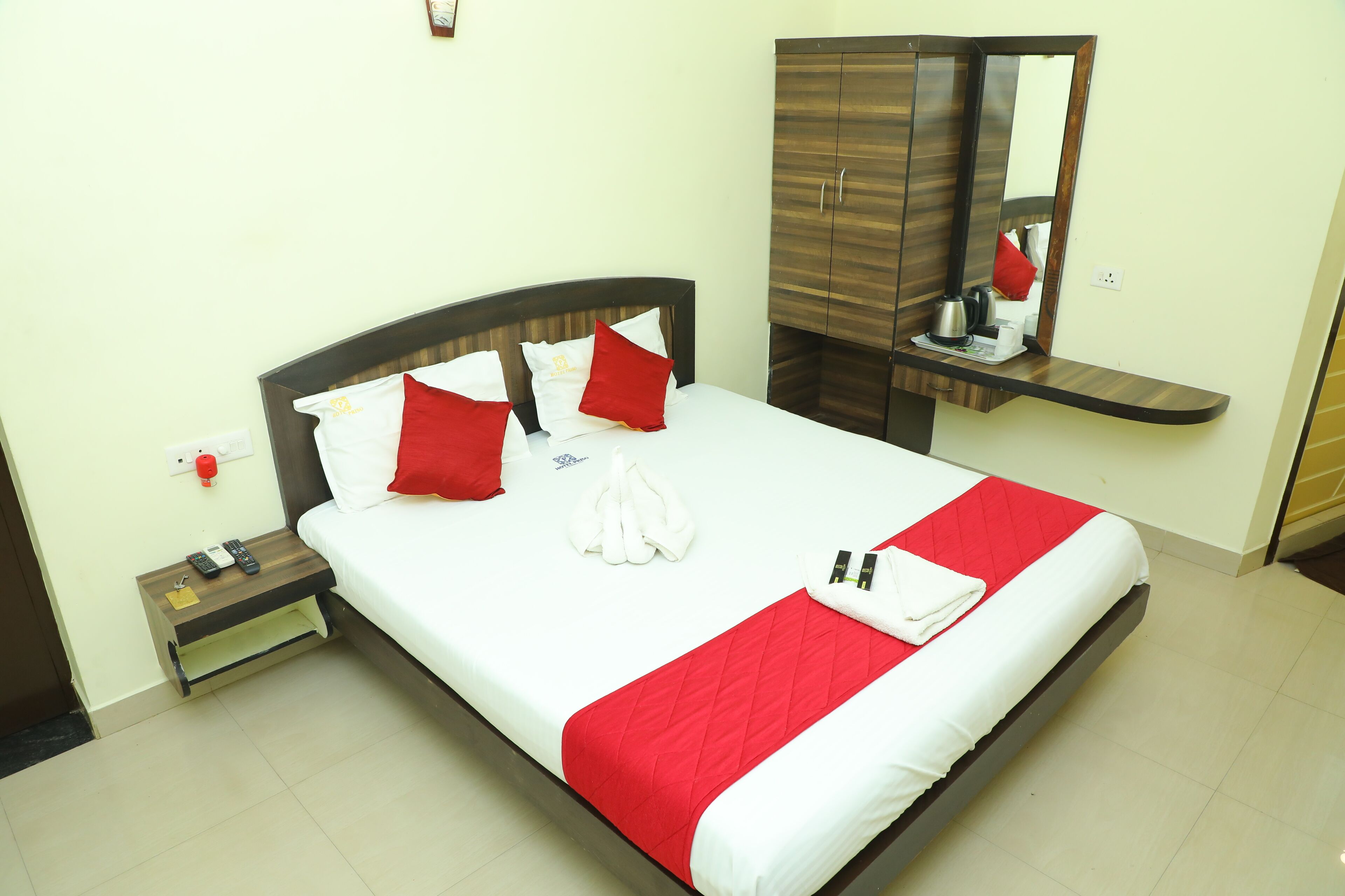 A neatly made bed with red and white bedding, a wooden nightstand with a remote control, and a mirror above the bed.