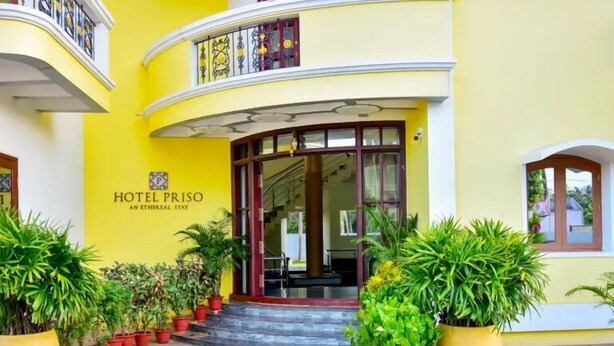 A hotel entrance with a yellow facade, a sign reading "HOTEL PRISO AN ETHEREAL STAY", and potted plants in front. - Hotel Priso (Puducherry)