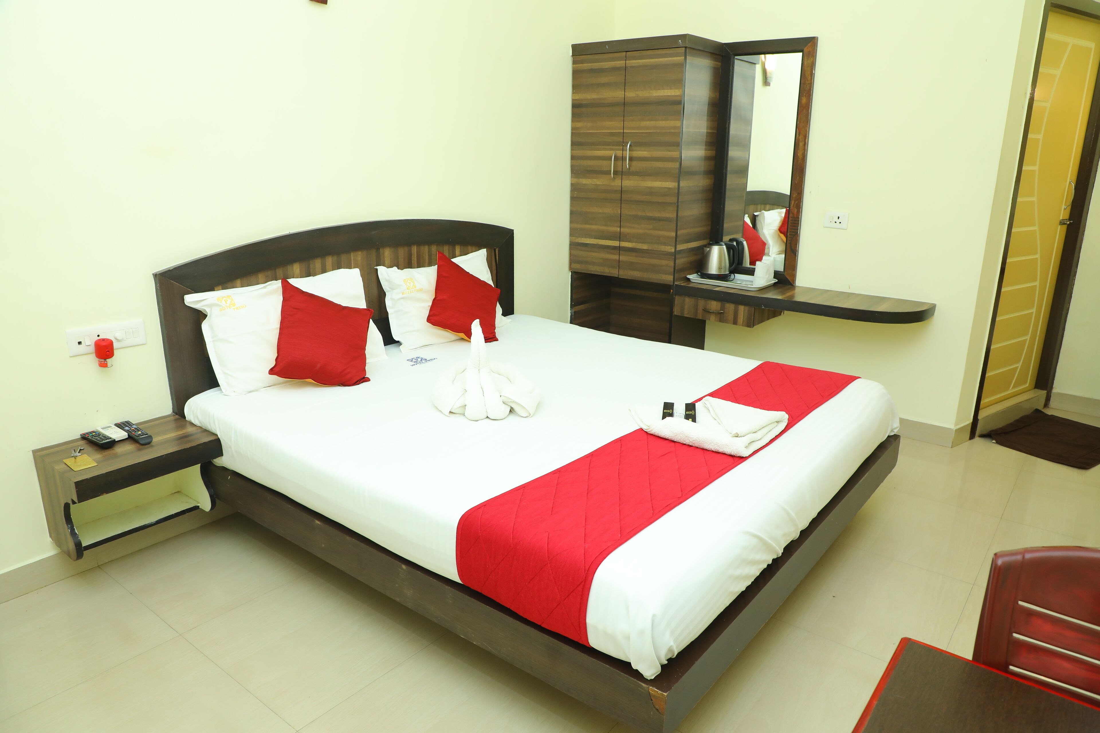 Ein ordentlich bezogenes Bett mit roter und weißer Bettwäsche, ein Nachttisch aus Holz mit Fernbedienung, ein Spiegel über dem Bett und ein roter Sessel im Zimmer.