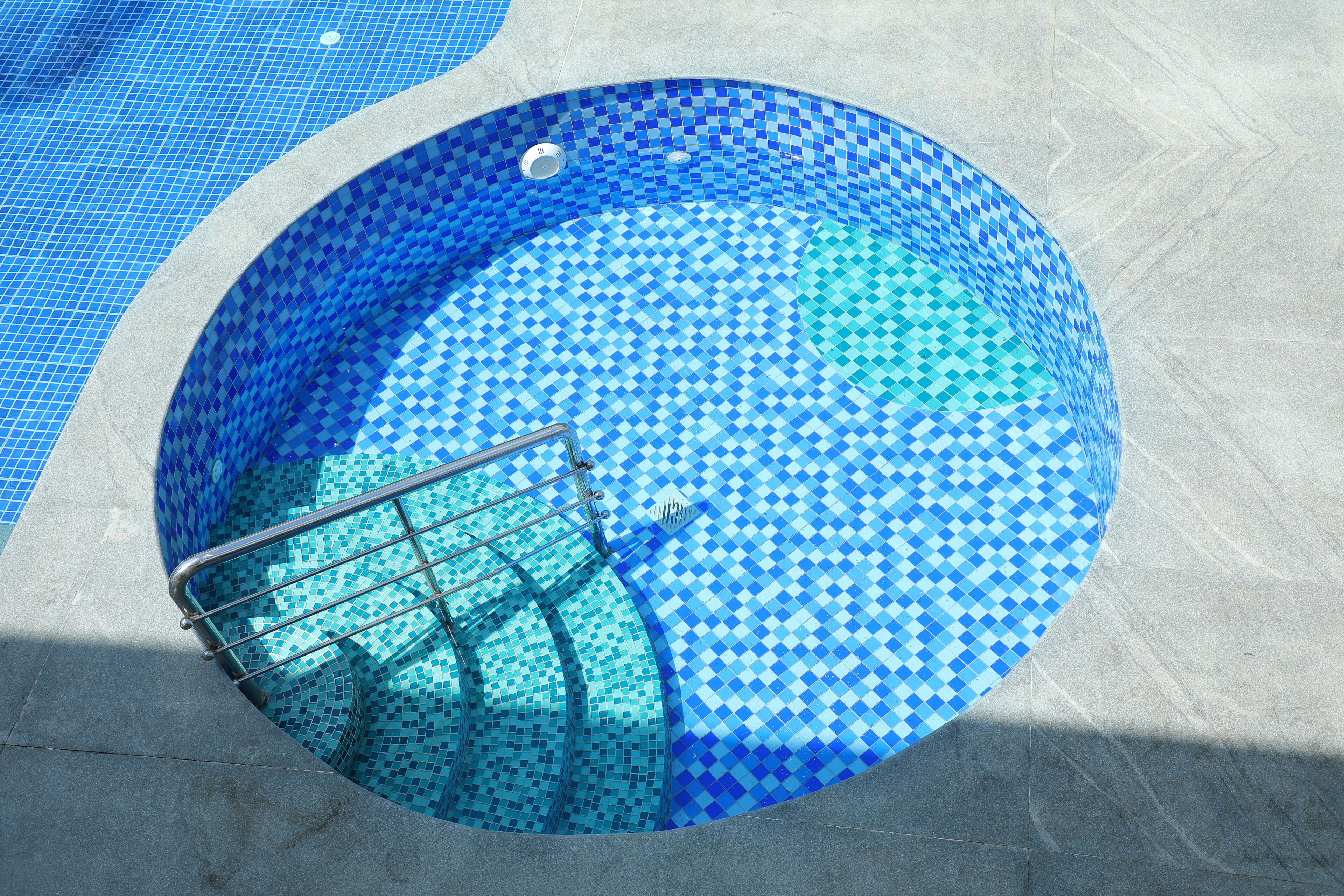 A circular pool with a blue mosaic tile pattern, a metal ladder, and a tiled deck surrounding it.
