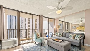 Un salon spacieux avec un grand canapé, une table basse et un fauteuil. Il y a un balcon avec vue sur de hauts immeubles.
