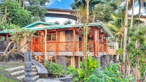 Una casa de madera con una escalera de piedra que lleva a un porche, rodeada de plantas tropicales y un camino de piedras.