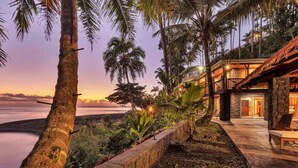 Una propiedad frente a la playa con un muro de contención de piedra, palmeras y una terraza de madera con un banco.