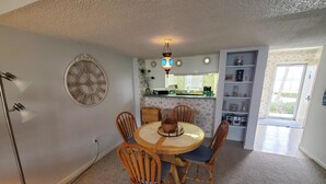 Un comedor con una mesa redonda, cuatro sillas de madera, un reloj de pared y una cocina visible a través de una puerta abierta.
