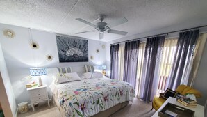 Un dormitorio con cama, mesitas de noche, un escritorio, ventilador de techo y cortinas.