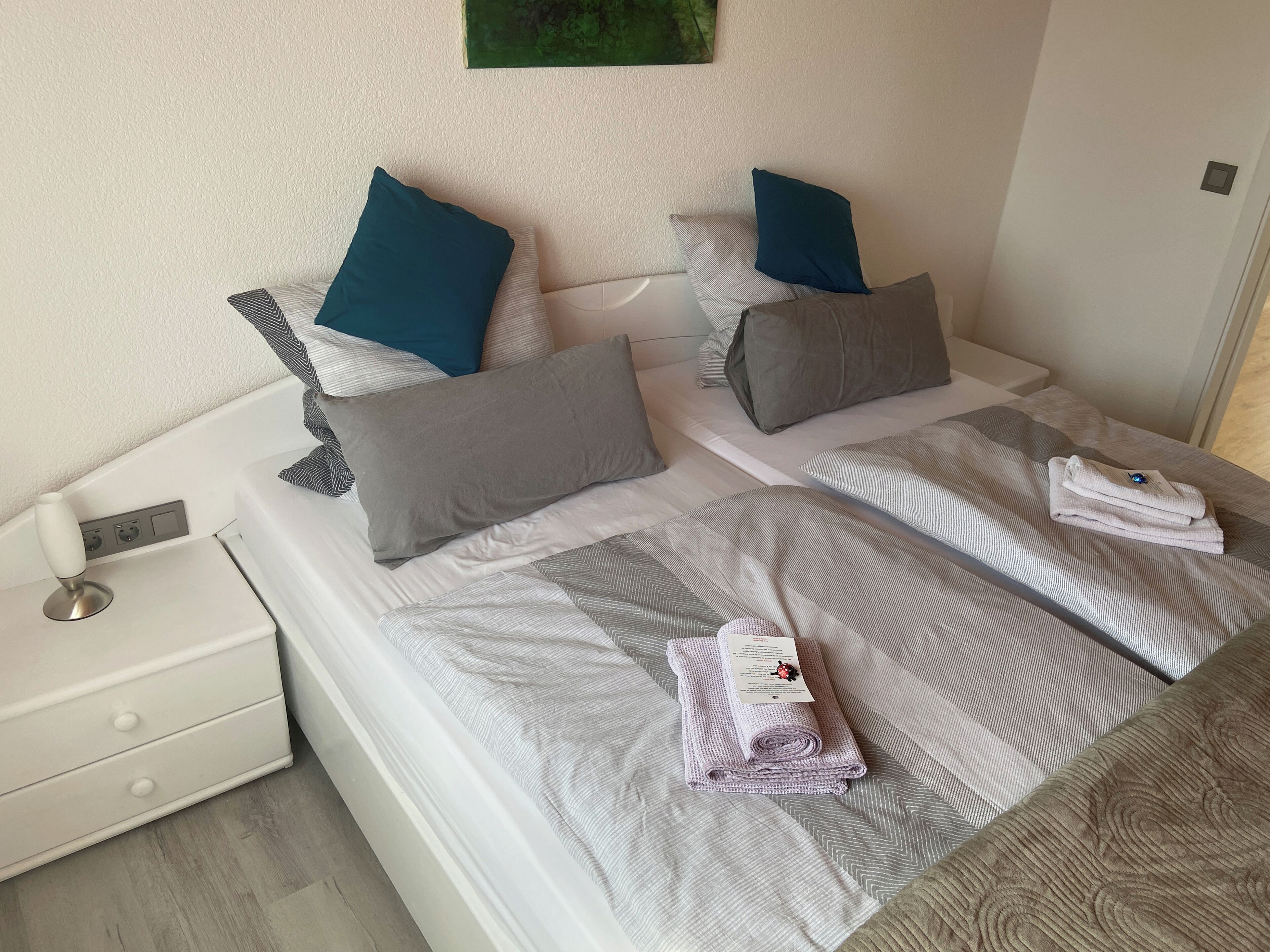 A hotel room with two beds, each with grey and blue pillows, a white bedside table, and a folded towel on the bed.