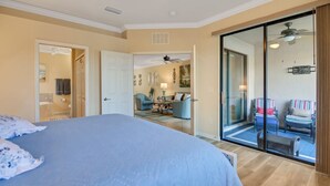A bedroom with a large bed, a sliding glass door leading to a balcony with seating, and a bathroom visible through an open door.