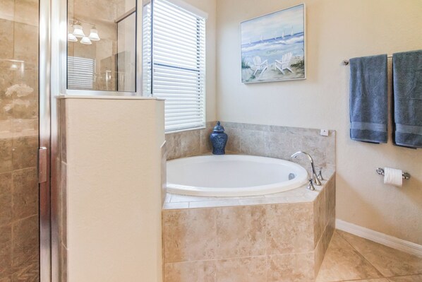 Une salle de bain avec une baignoire, un lavabo, une fenêtre avec des stores, un vase et deux serviettes sur un porte-serviettes.