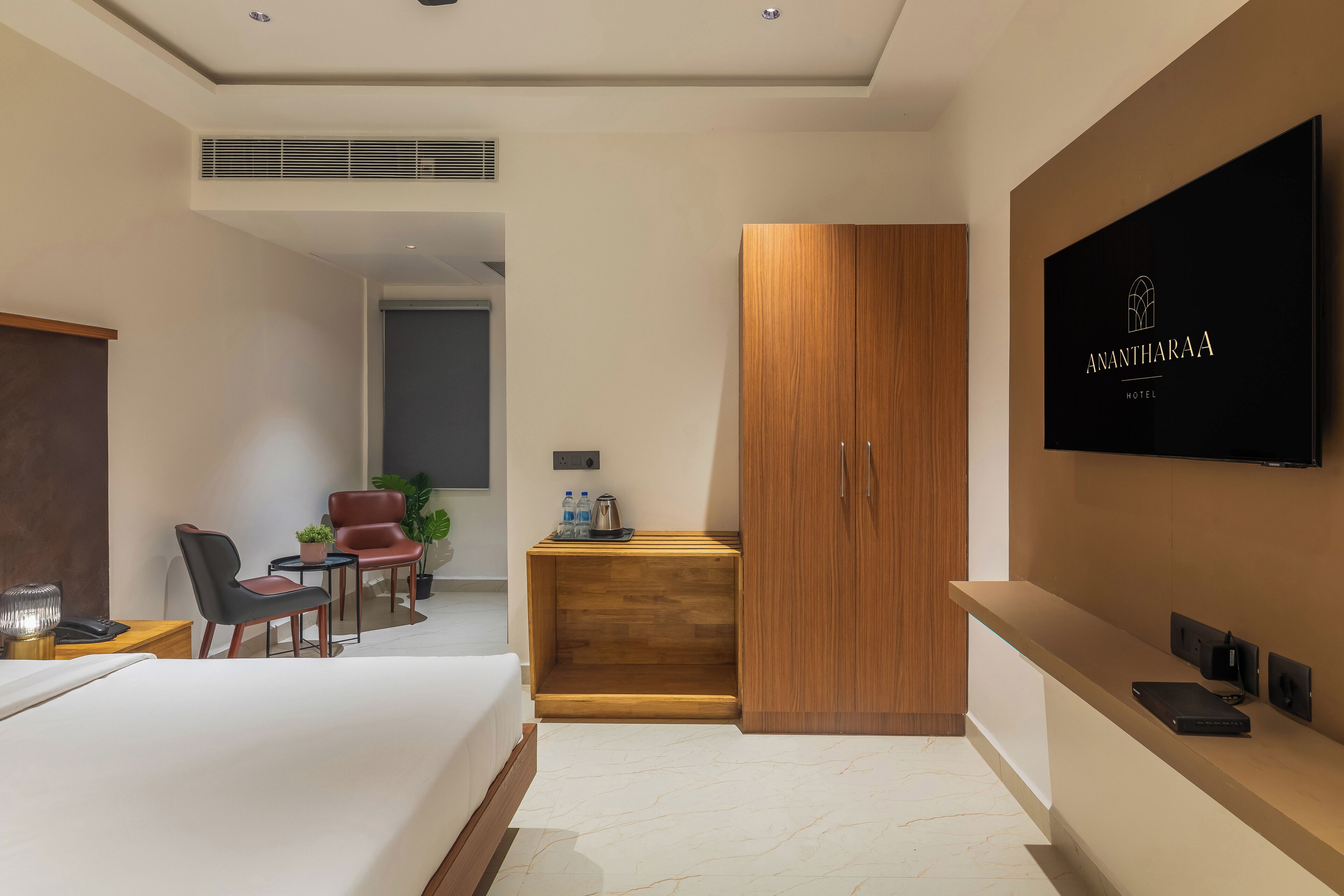 A modern hotel room with a large bed, a wooden wardrobe, a desk with a plant, and a TV mounted on the wall.
