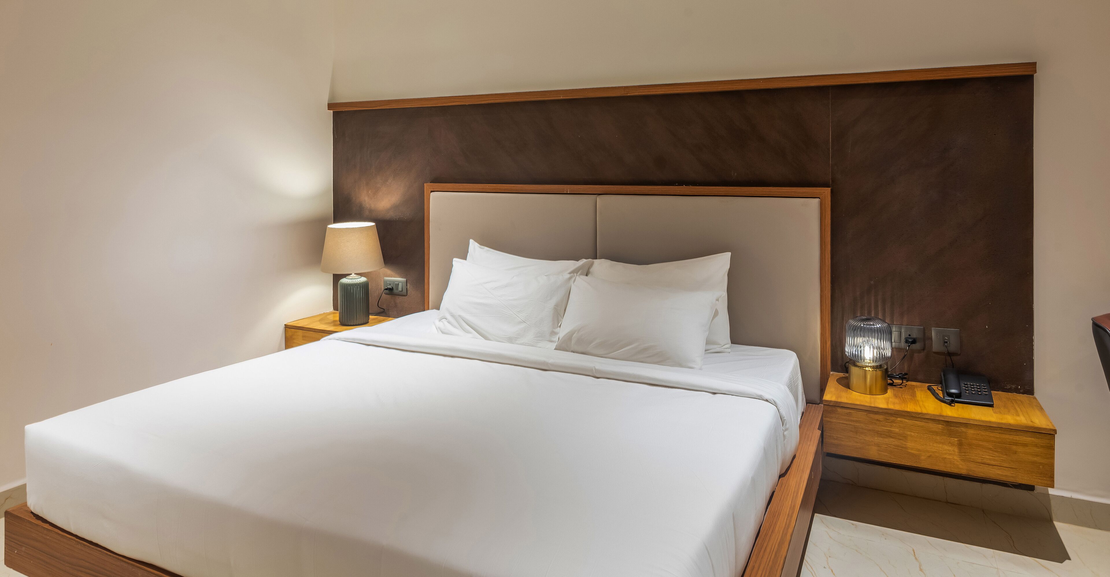 Un lit bien fait, avec du linge de lit blanc, une tête de lit en bois et une table de chevet sur laquelle se trouvent une lampe et un téléphone.
