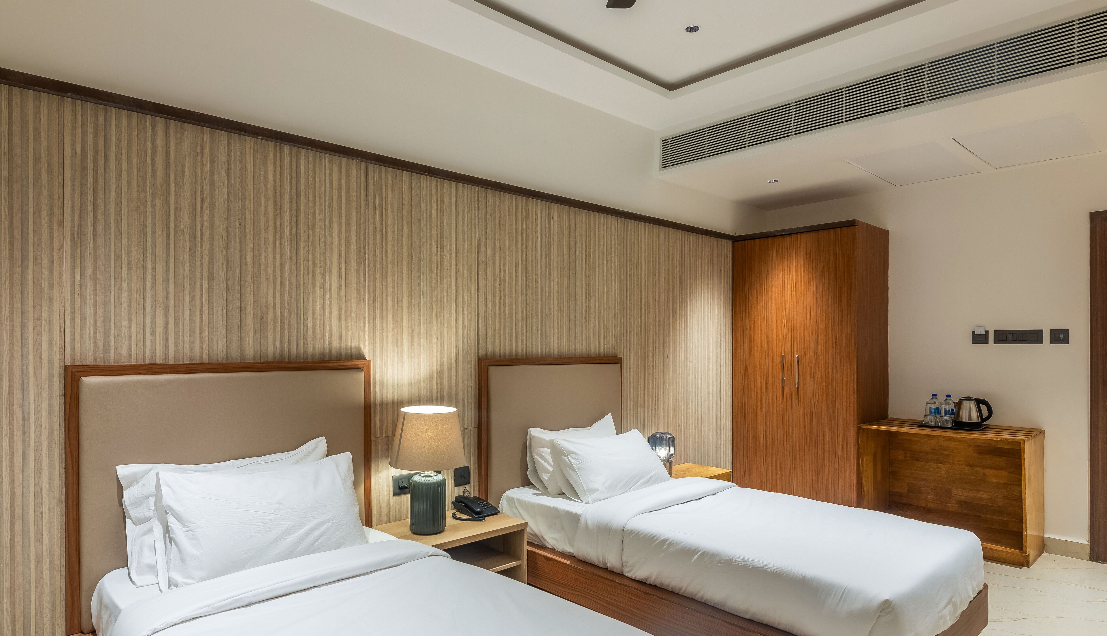 A hotel room with two single beds, a wooden wardrobe, a bedside table with a lamp, and a small side table with a kettle and water bottles.
