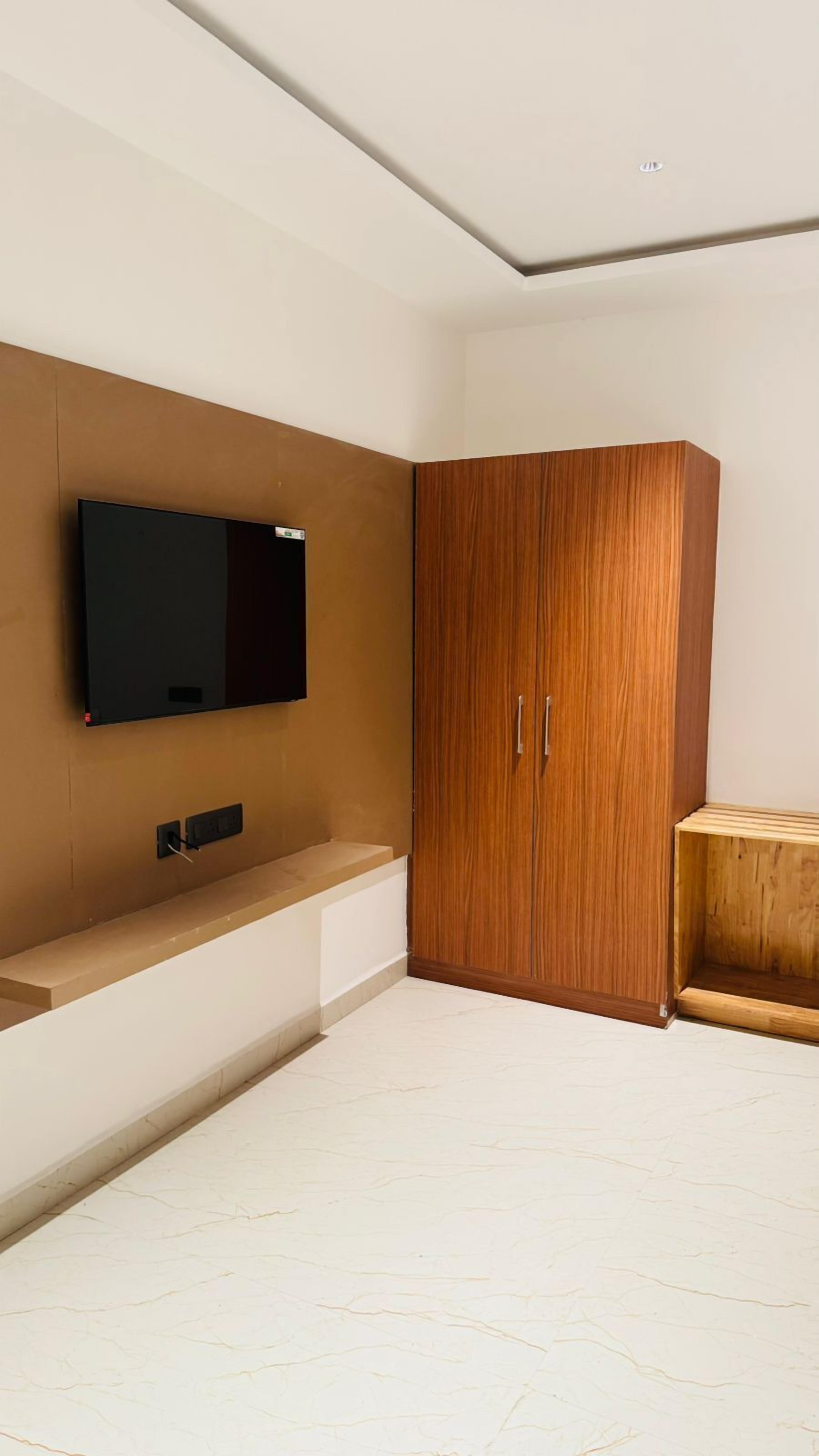 A room with a flat-screen TV mounted on the wall, a wooden wardrobe with two doors, and a small wooden table.