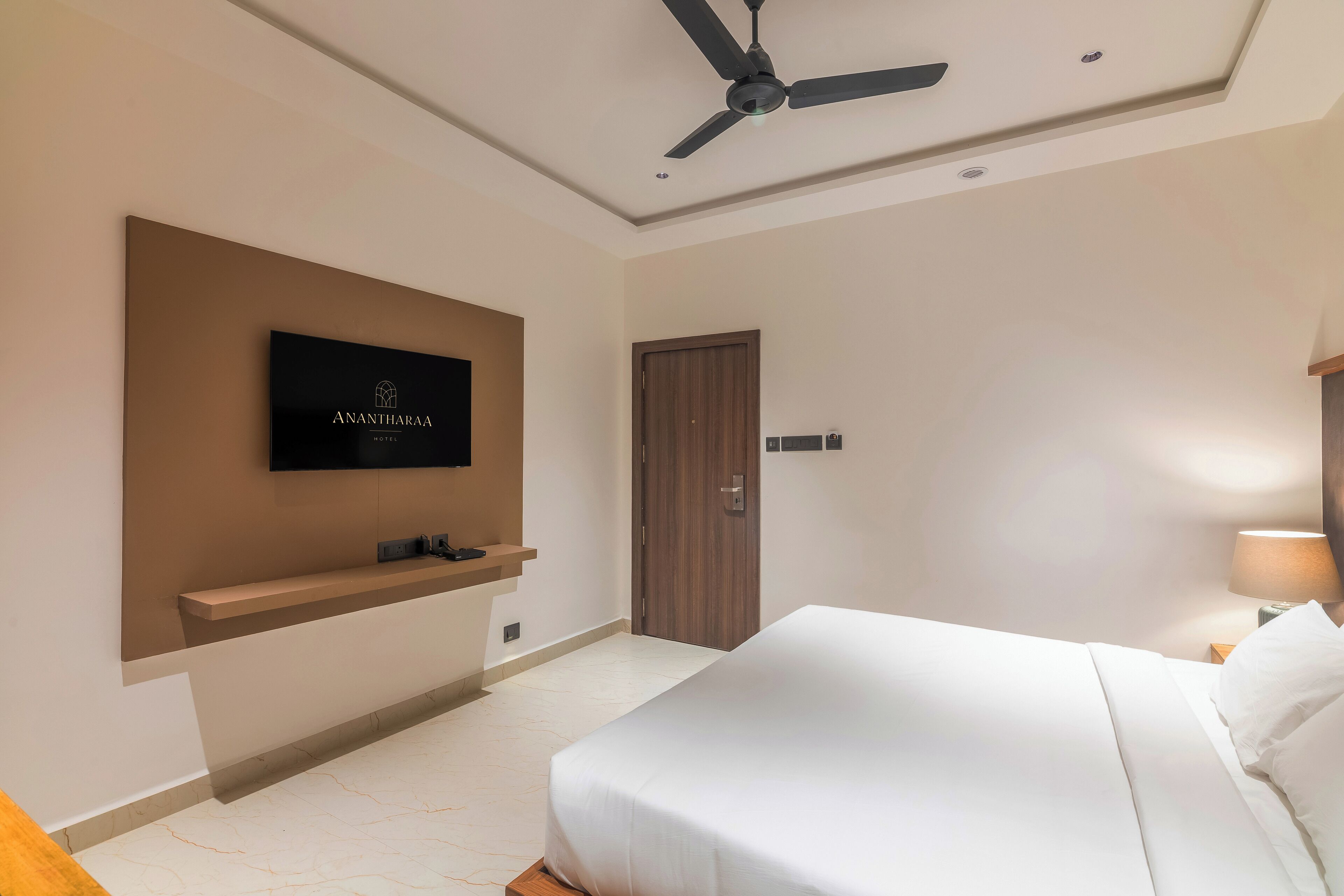 A modern hotel room with a large bed, a mounted TV, a ceiling fan, and a wooden door.