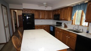 Uma cozinha com armários de madeira, bancada de mármore branco, frigorífico preto, micro-ondas, fogão e máquina de lavar loiça.