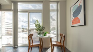 Una habitación moderna con ventanas amplias, una mesa redonda con sillas, una planta en maceta y un cuadro abstracto enmarcado.