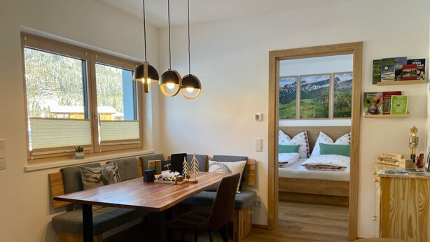 A modern living room with a wooden dining table, a sofa, and a view of a mountain landscape through a window. - Beagzeit Ferienwohnung (Lofer)