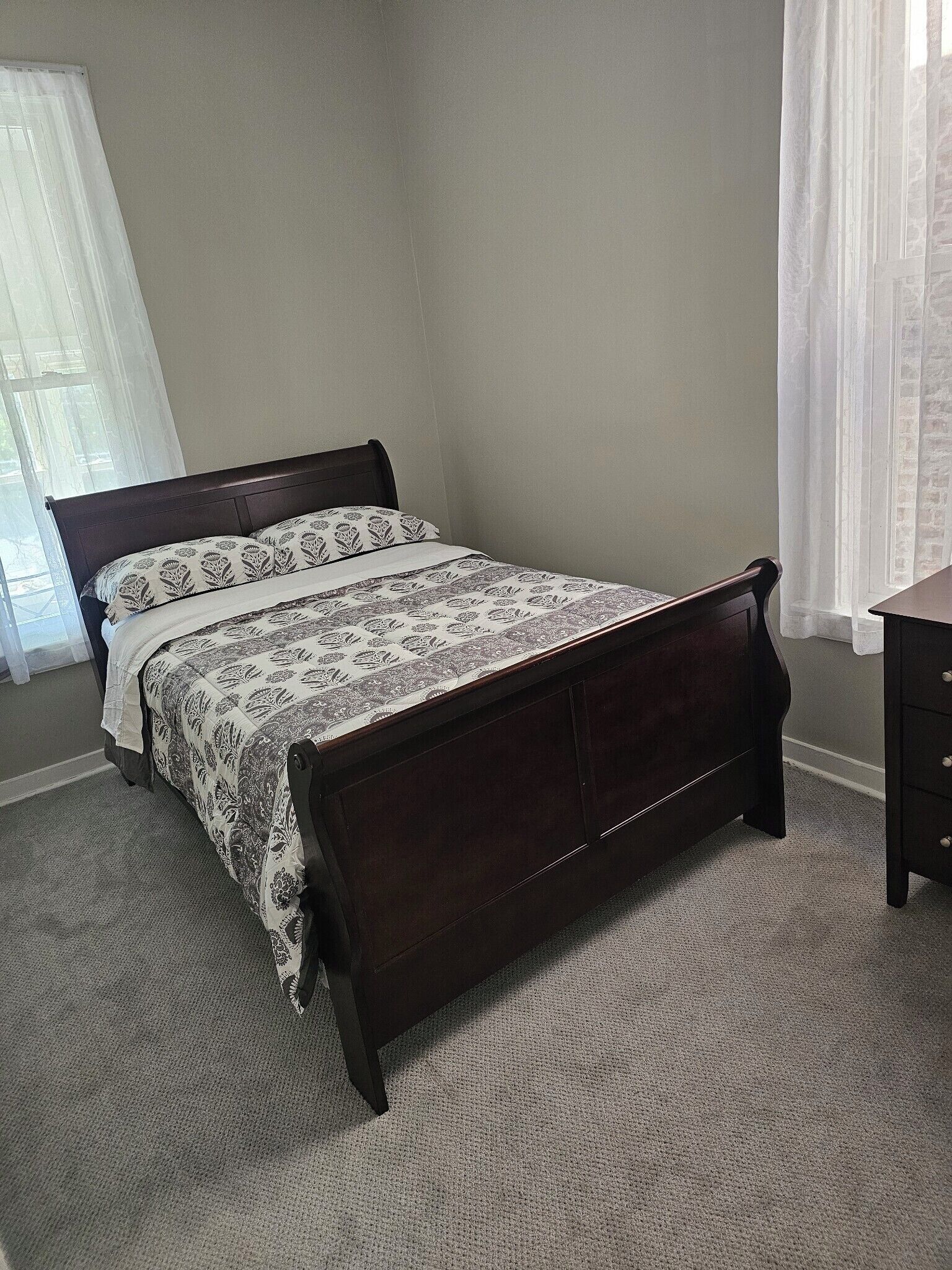 A bedroom with a dark wooden bed, patterned bedding, a small dresser, and a window with sheer curtains.